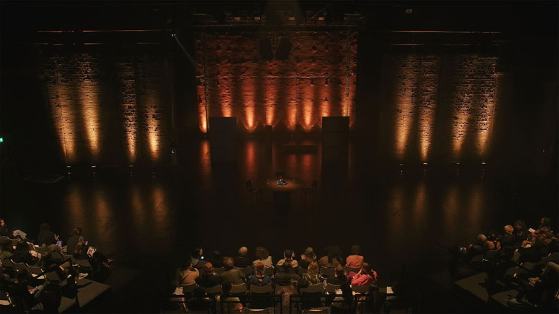 Audience seated before the orange-lit Tramway stage, the round table small in the distance