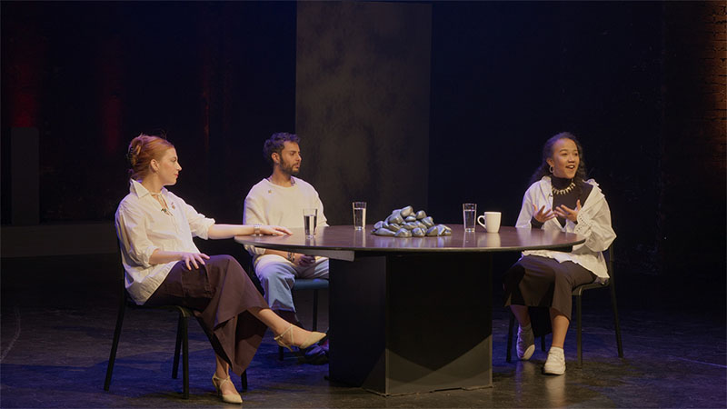 Three performers in white shirts at the round table, one gesturing as she speaks