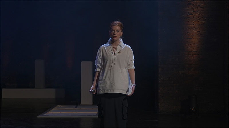 Red-haired performer in a white shirt stands centre stage, brick wall lit warmly behind