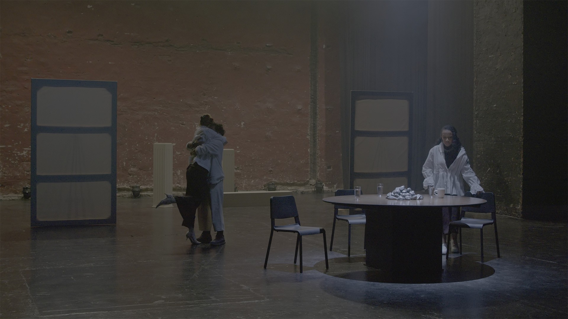 Video documentation still: two performers embrace while a third stands at a round table on a hazy stage