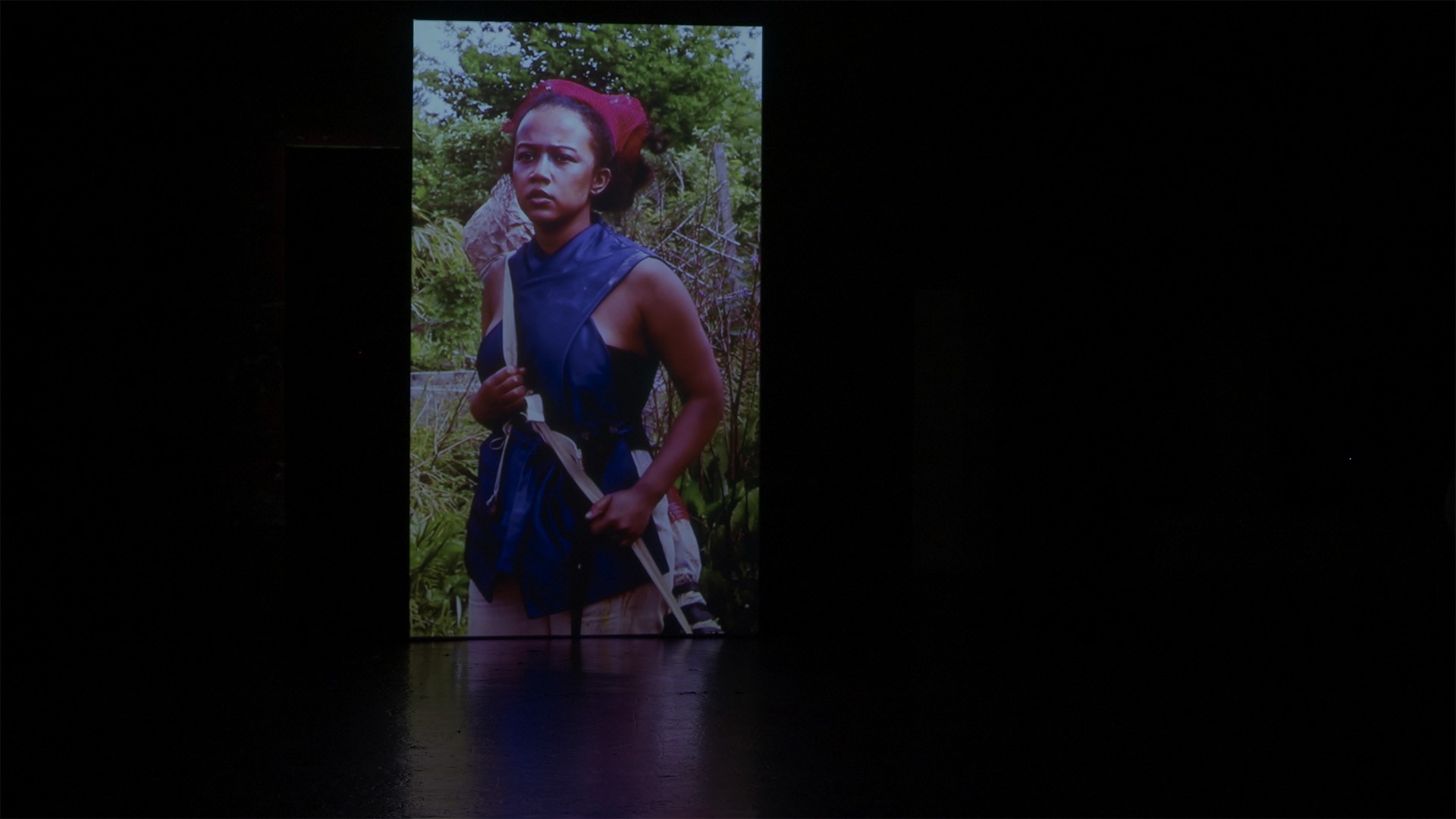 Projected film of a woman in a red headwrap and blue garment amid greenery glows in the dark theatre