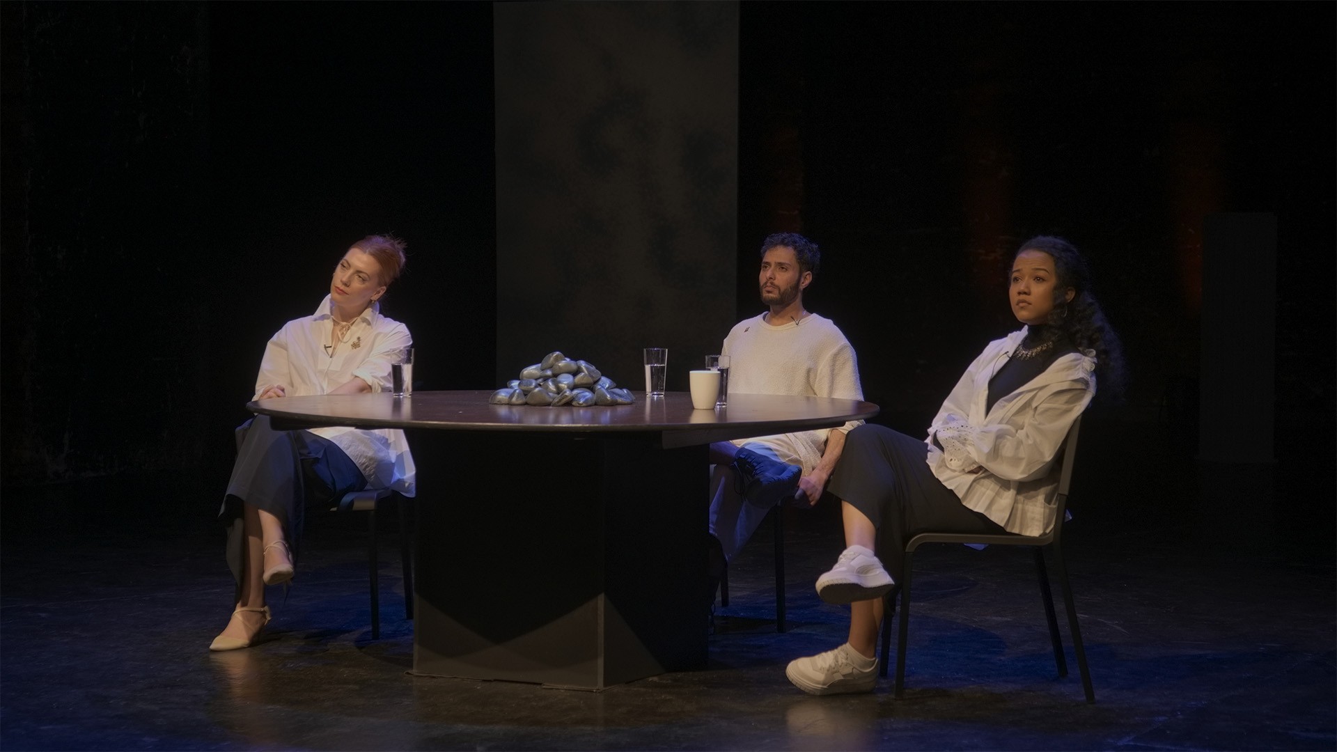 Three performers lean back from the round table, gazing outward, stones piled between glasses and a mug