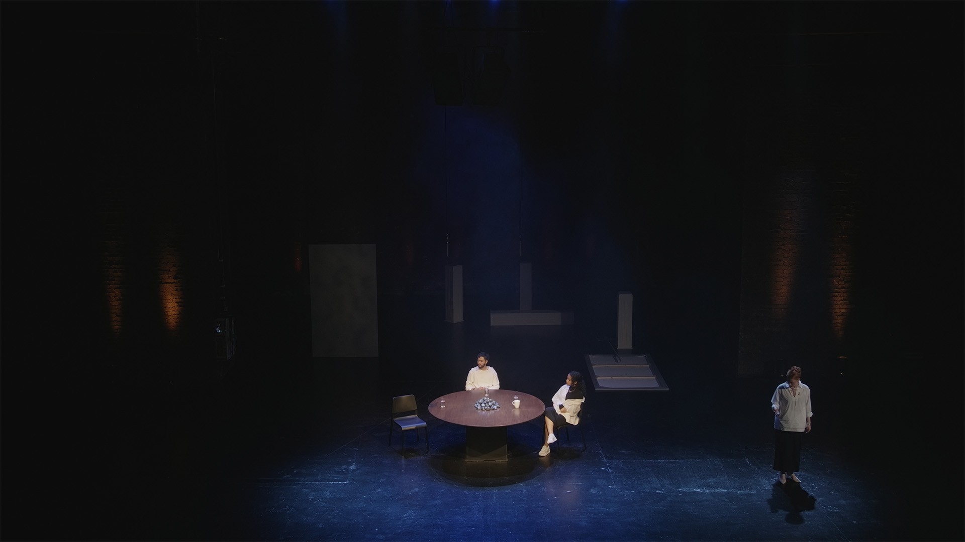 High wide video still: two performers sit at the round table while a third stands apart in dim blue light