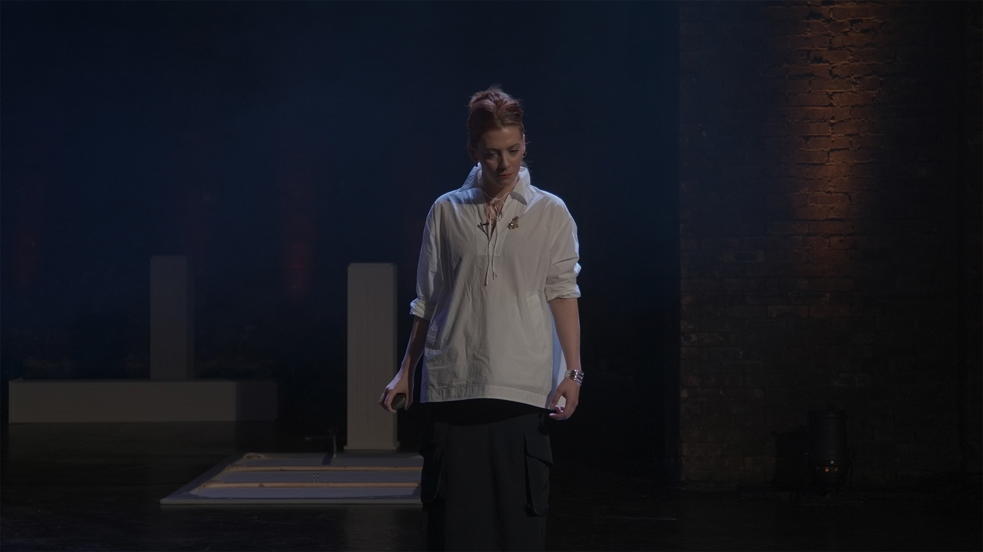 Performer in a loose white shirt looks down, lit against a brick wall on the darkened stage
