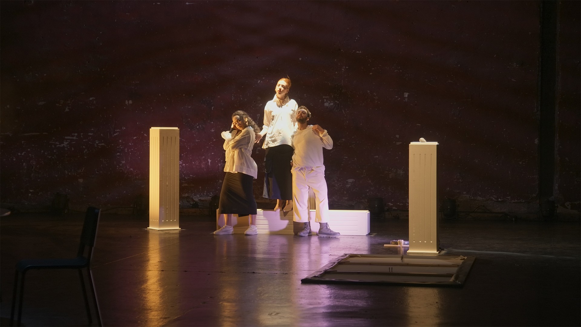 Three performers pose close together among glowing white plinths under warm light