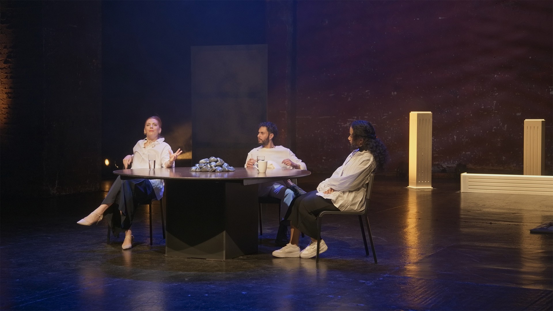 Three performers talk at the round table, warm-lit brick wall and glowing plinths behind