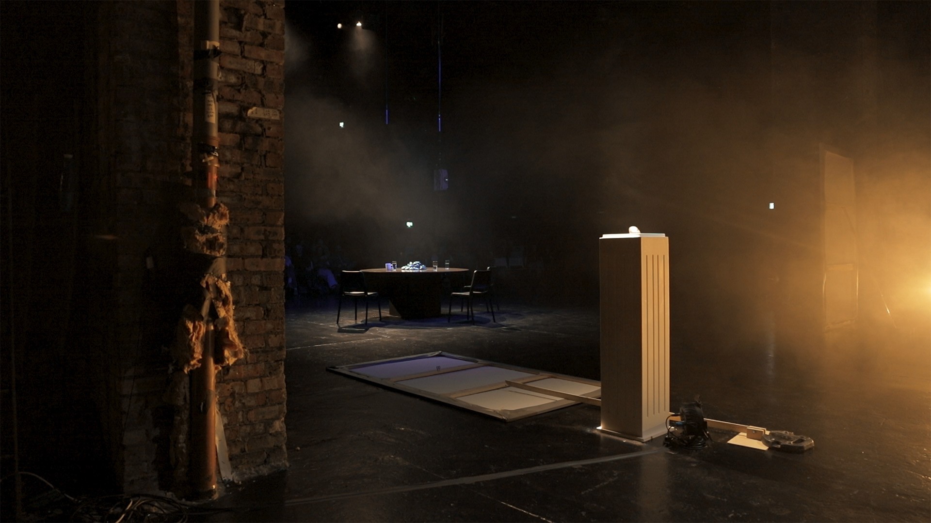 View from the wings past a brick column toward the round-table set, a lit plinth standing in haze