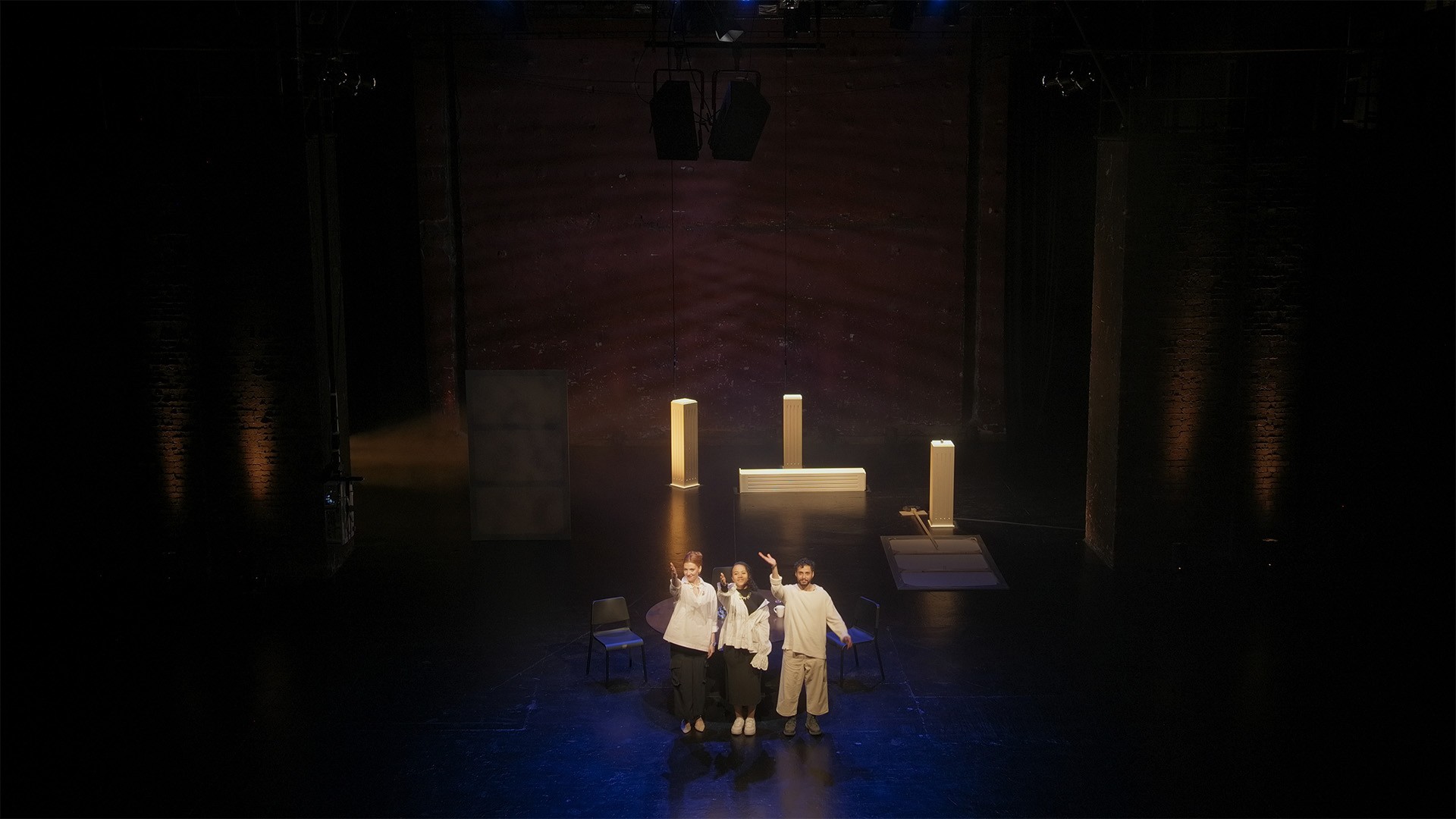 Seen from above, three performers wave at the curtain call beside glowing white plinths