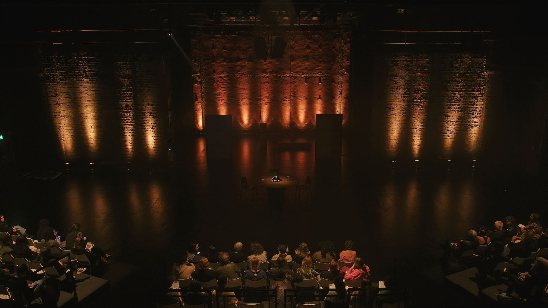 Audience watch the distant round-table set at Tramway, brick walls washed in orange uplight