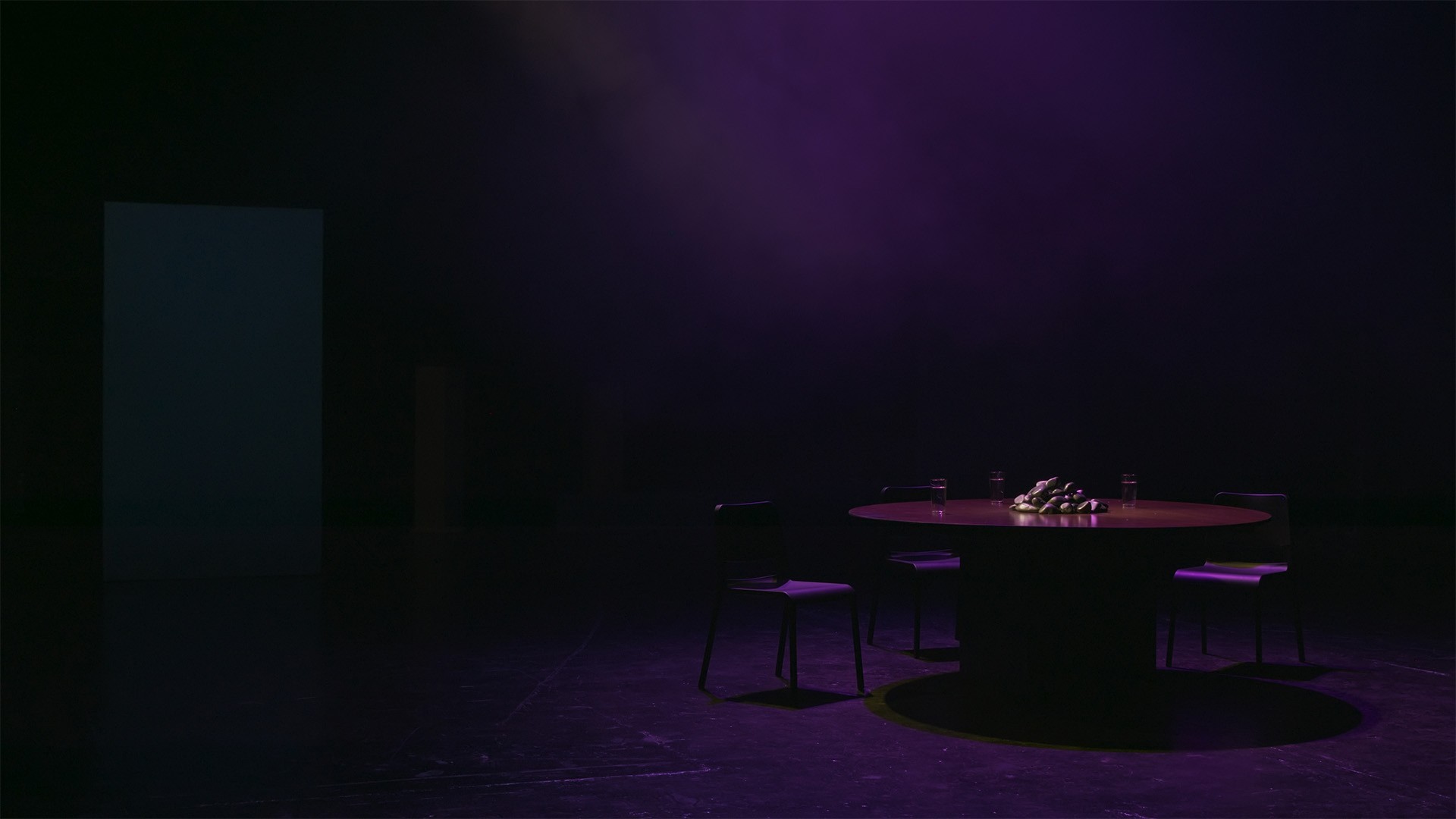 Empty set in deep purple light: a round table holding a pile of stones, ringed by three chairs