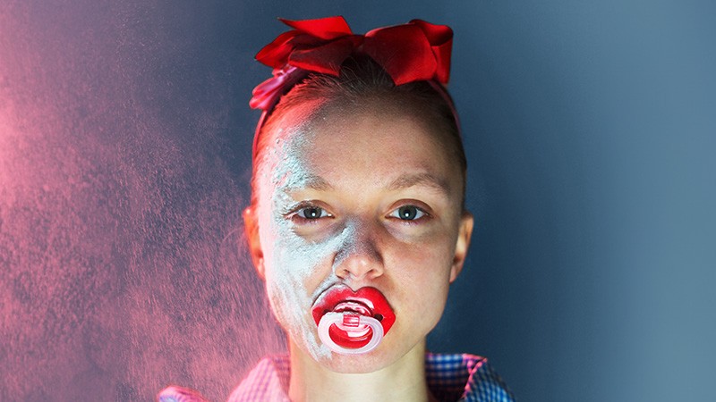 Publicity image: performer with red hair bow and red dummy in mouth, face dusted with powder