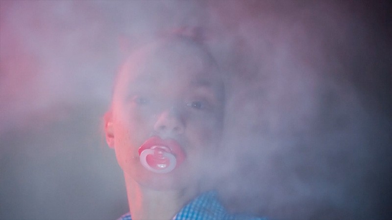 Trailer still of a face with a red dummy in mouth emerging through pink-tinted haze