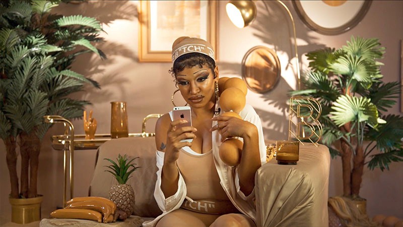 Film still of Nima Séne as Beige B*tch scrolling a phone in an all-beige lounge with palms and a pineapple