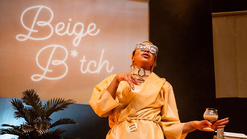 Nima Séne in a beige latex robe holds a glass, neon Beige B*tch script glowing on the screen behind