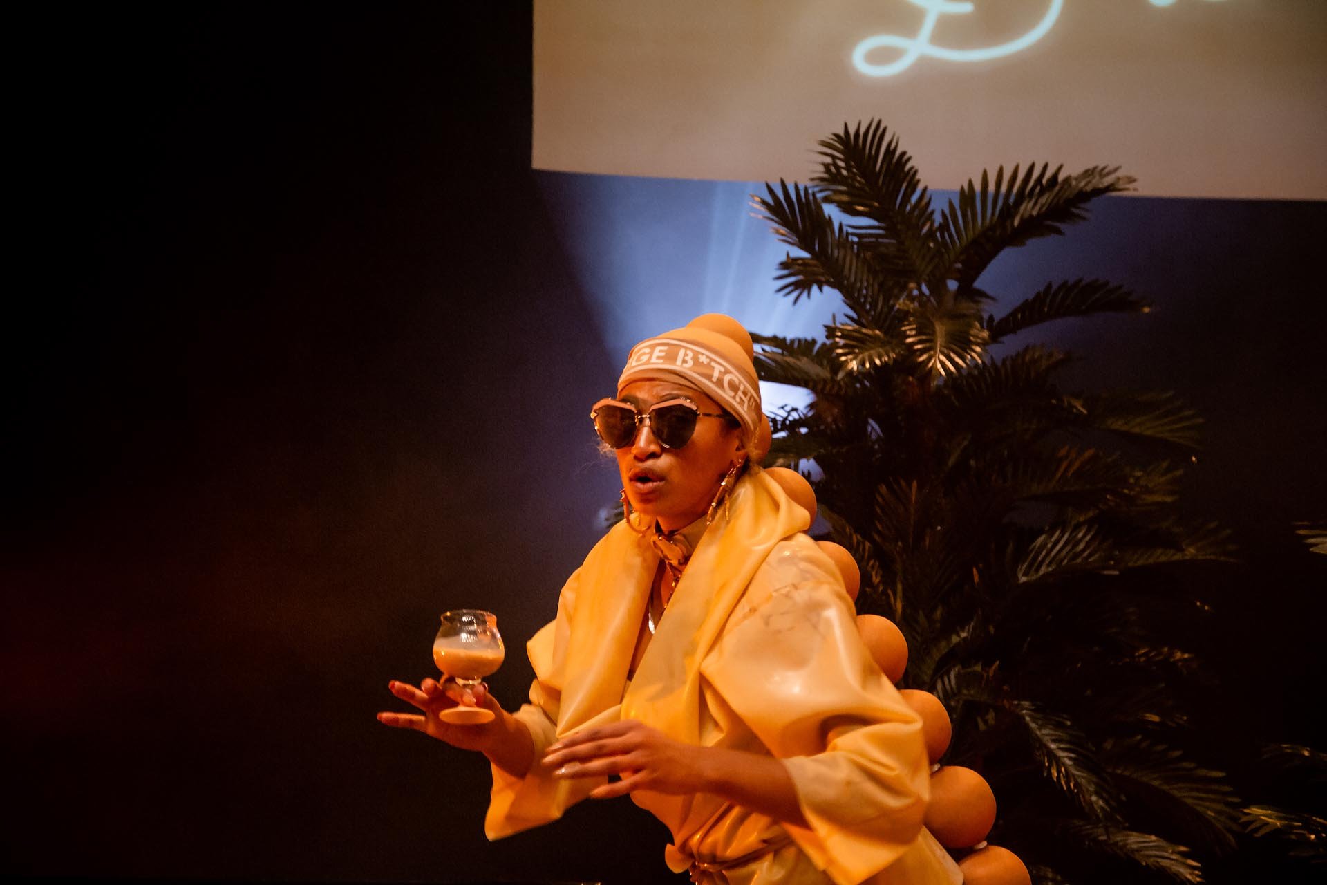Nima Séne in yellow latex and Beige B*tch headband holds a creamy drink on stage at Take Me Somewhere