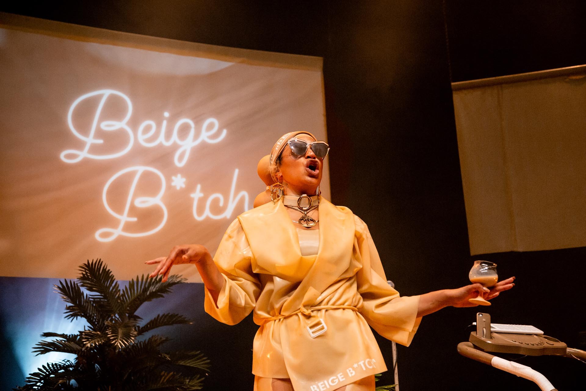 Nima Séne gestures mid-speech with a drink in hand, neon Beige B*tch script projected behind her