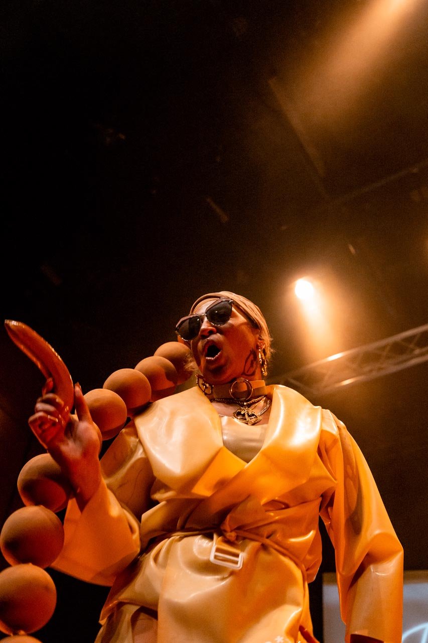 Low-angle performance photo of Nima Séne calling out, sausage prop in hand, draped in giant beads
