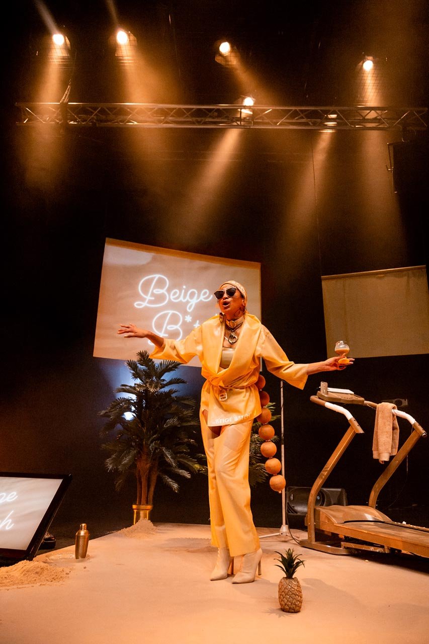 Nima Séne performs arms spread on a sandy white stage with palm tree, gold treadmill and pineapple