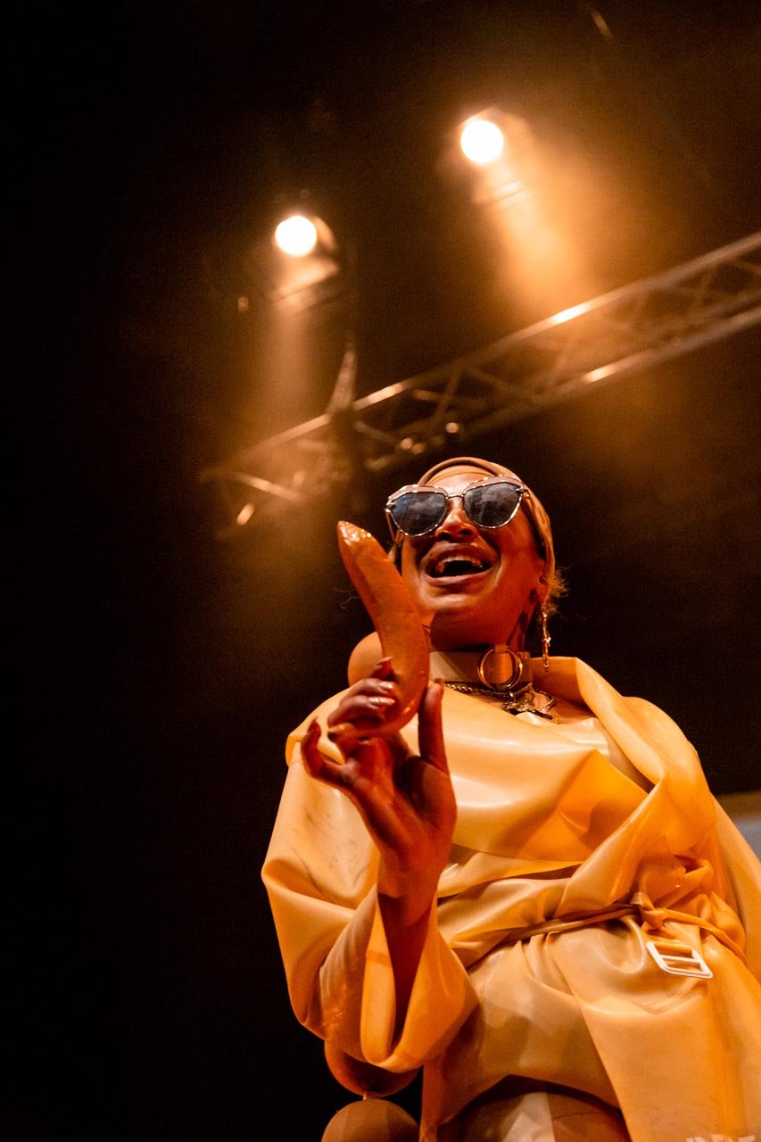 Nima Séne laughs under stage lights, brandishing a sausage prop in a beige latex coat