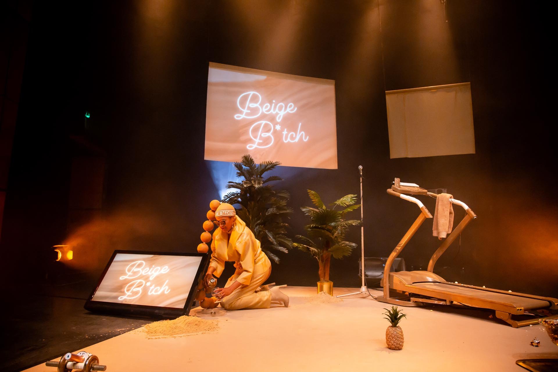 Nima Séne kneels on a white stage among palm trees, a gold treadmill and neon Beige B*tch projections