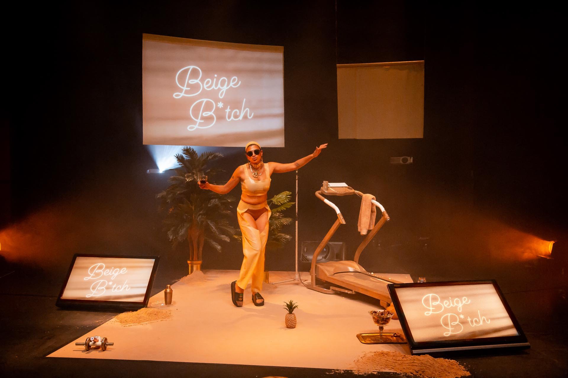 Nima Séne stands with arms outstretched between a palm tree and treadmill under Beige B*tch projections