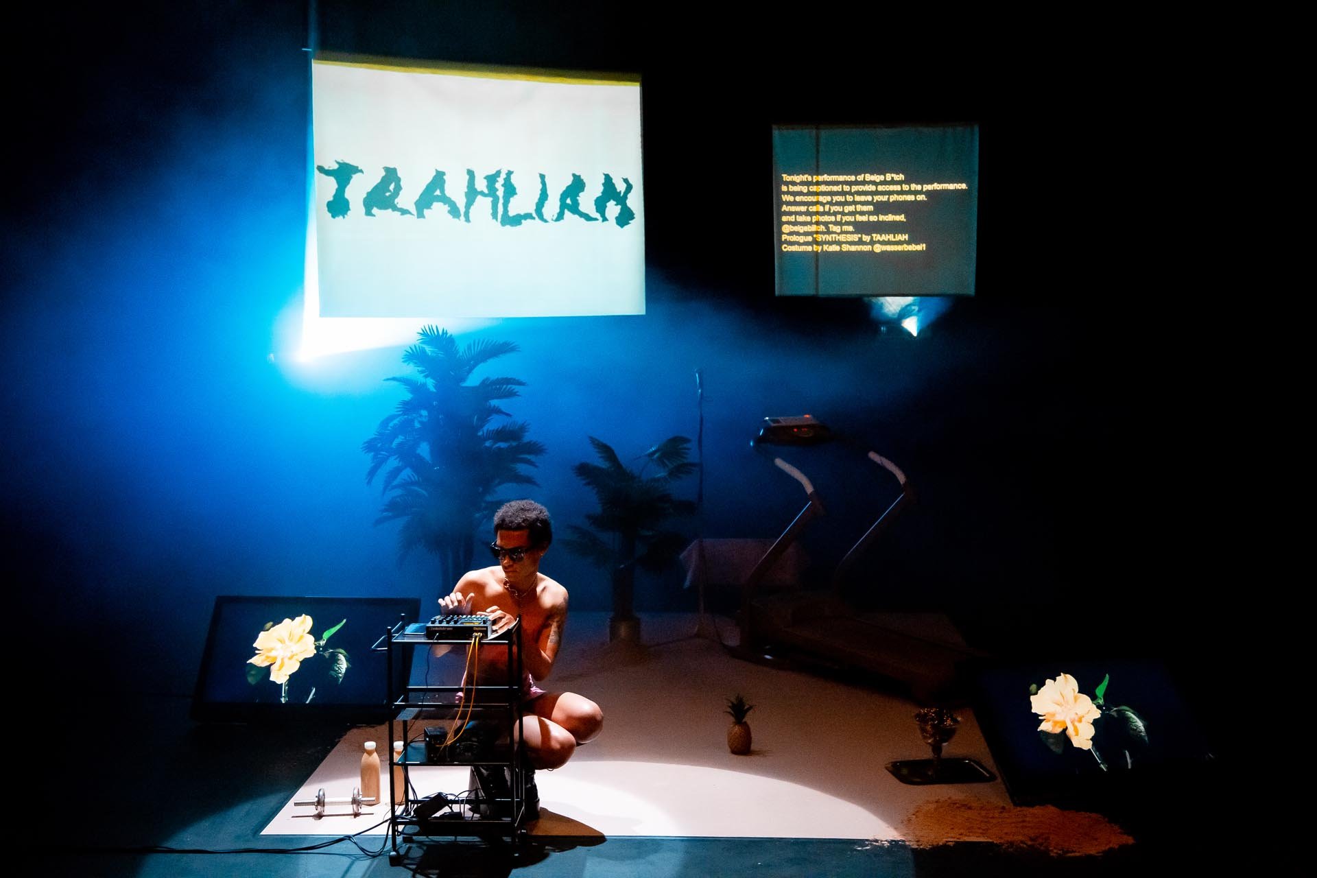 TAAHLIAH crouches over a synthesiser in blue haze, the name TAAHLIAH projected on a screen above