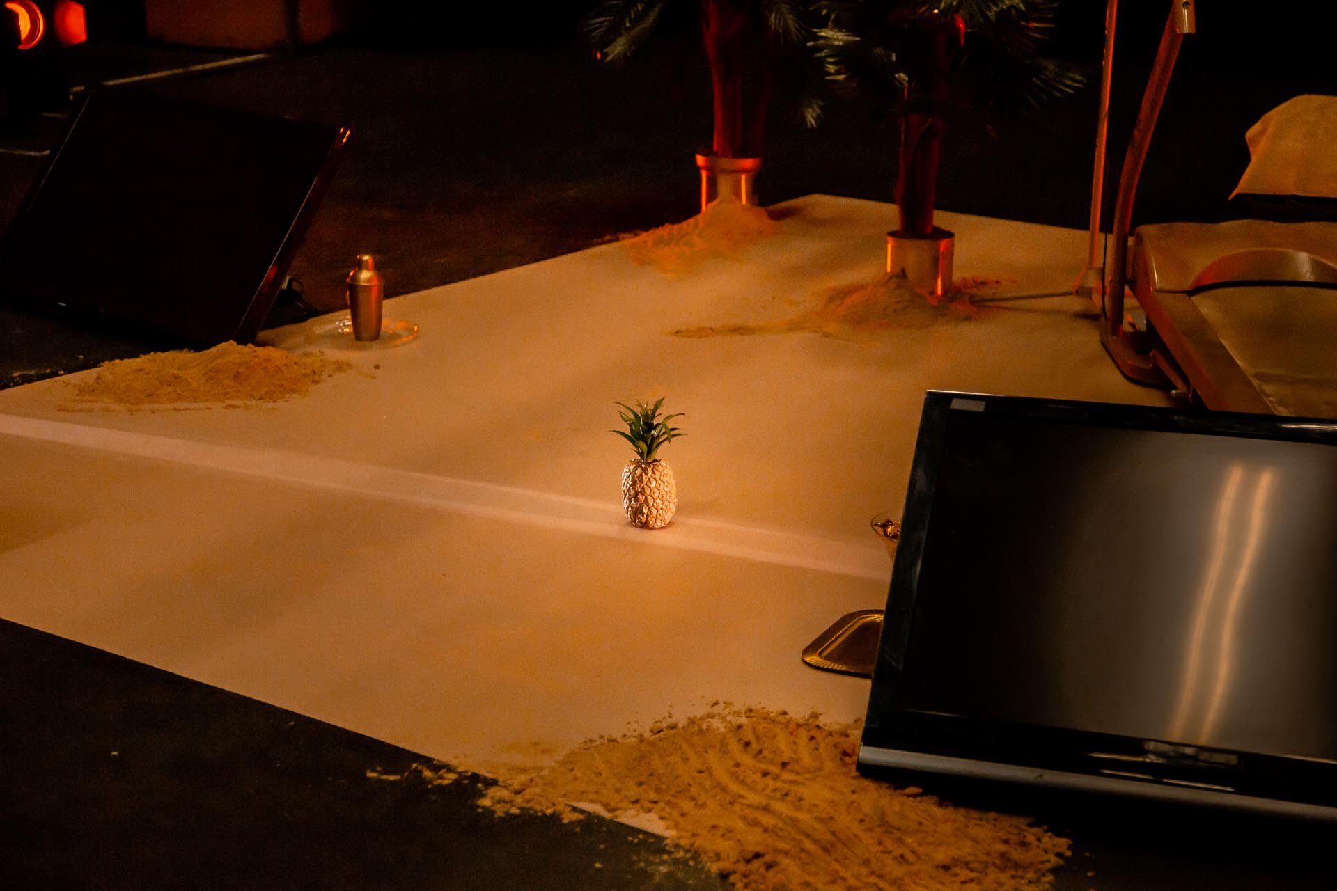 Set detail: a miniature pineapple on a white floor scattered with sand, beside a TV and treadmill