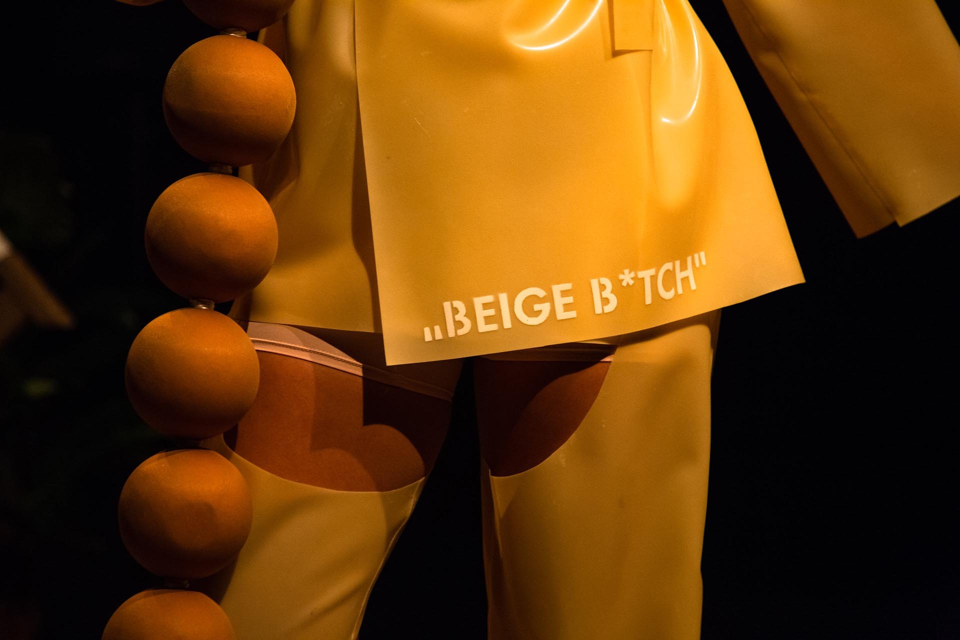 Costume detail: yellow latex printed with Beige B*tch beside a strand of giant wooden beads