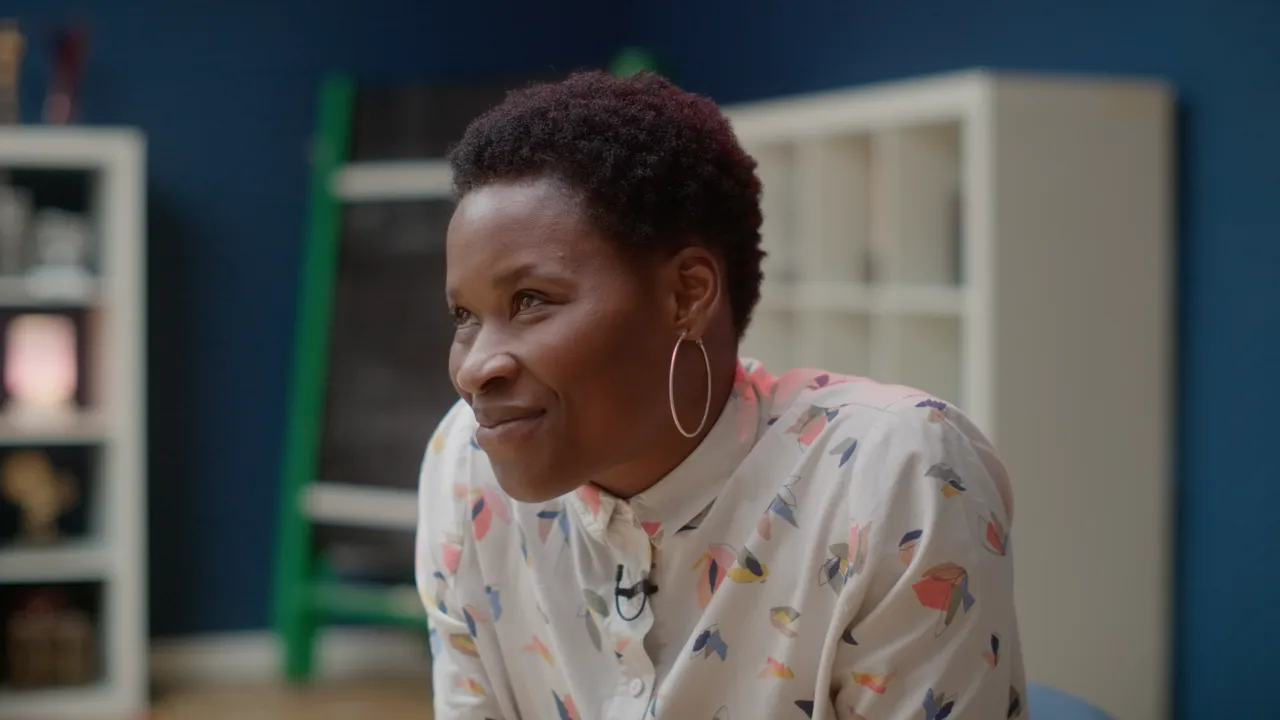 Adura Onashile, wearing a lavalier microphone, smiles in a documentary-style film still