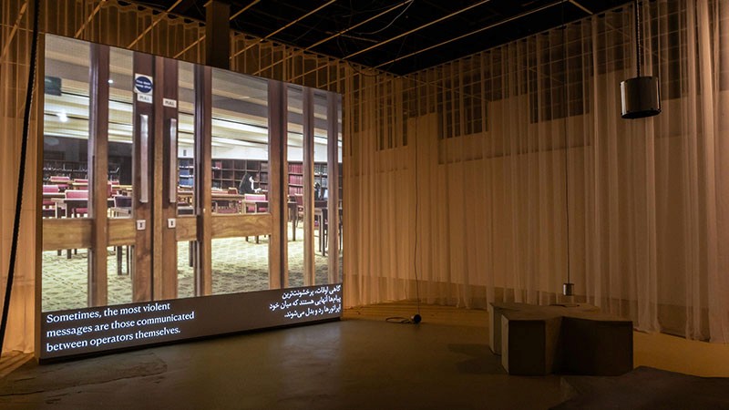 Installation view: large projection screen showing library footage in a gallery space hung with sheer curtains