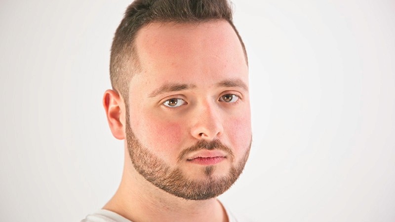 Trailer still: close-up of a bearded performer gazing deadpan at the camera against a white backdrop.