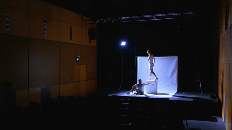 Two performers pose on a small white stage set in a dark theatre, one standing on a plinth, one seated below.