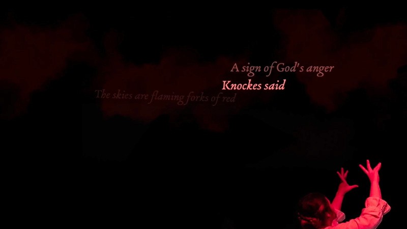 Red-lit captions reading 'A sign of God's anger, Knockes said' above a performer with raised hands; simulation still.