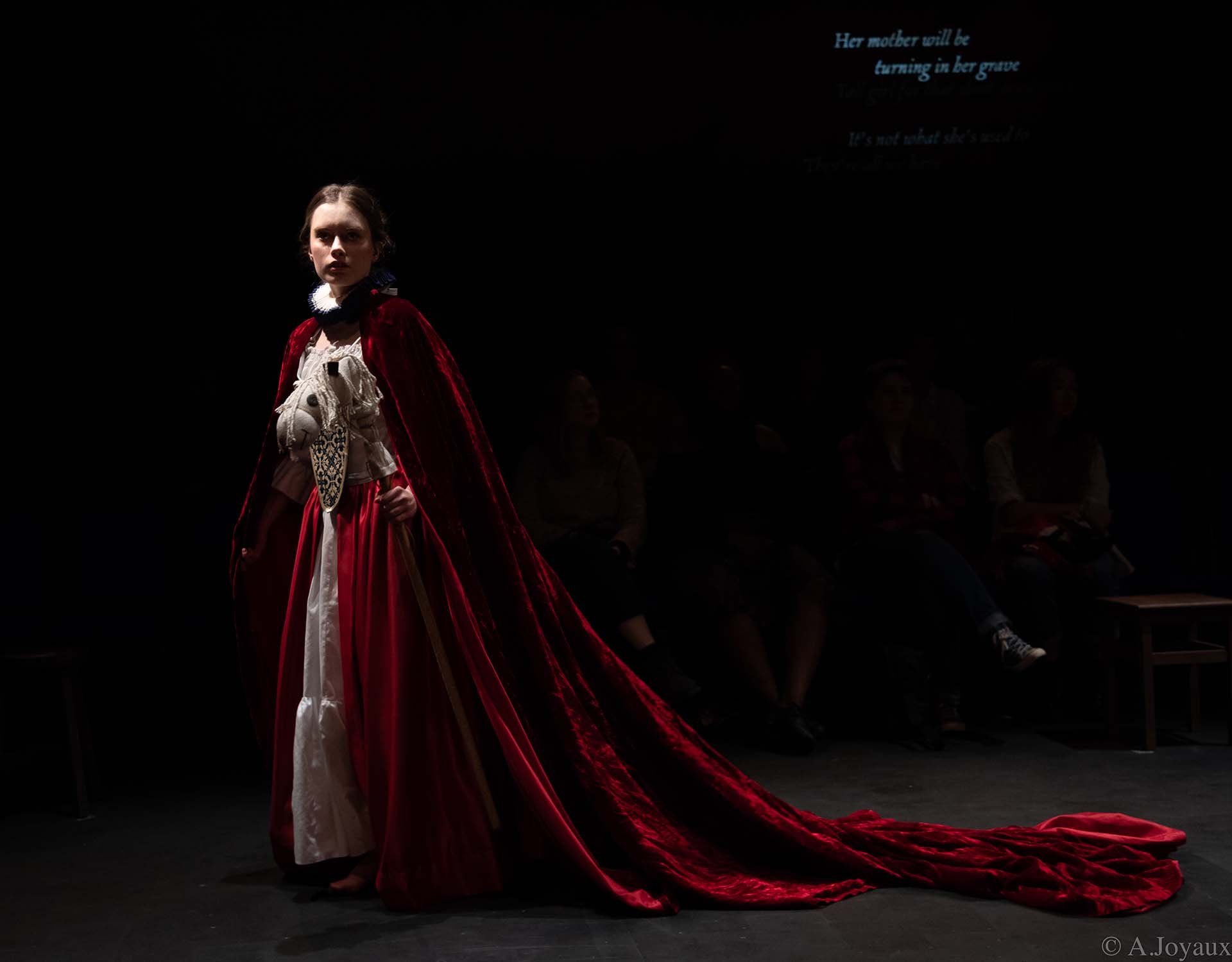Claire Wetherall as Mary in a trailing red velvet cloak, blue handwritten captions projected behind her.