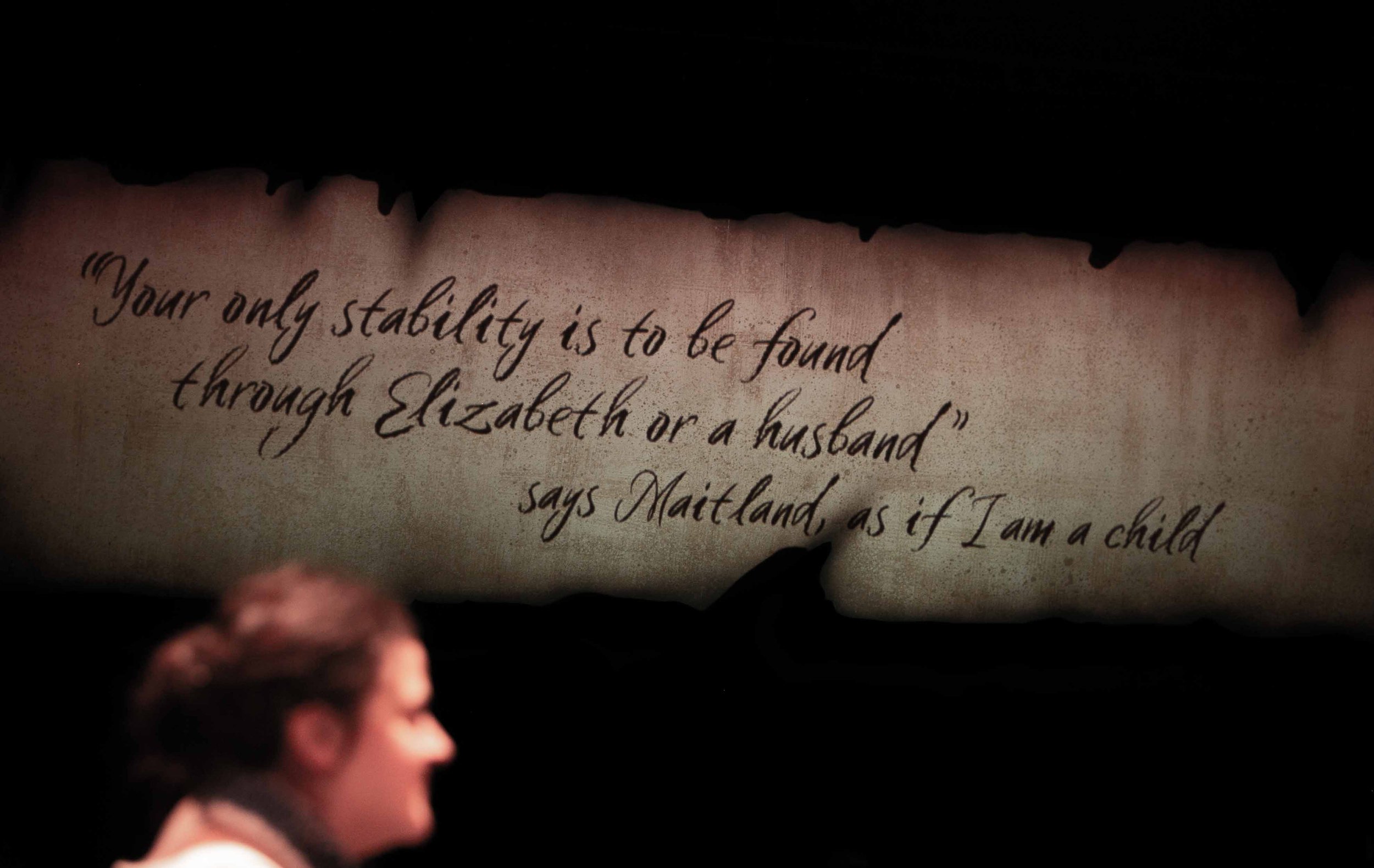 Captioning screen detail: a handwritten quote projected on torn parchment above a performer's silhouetted head.