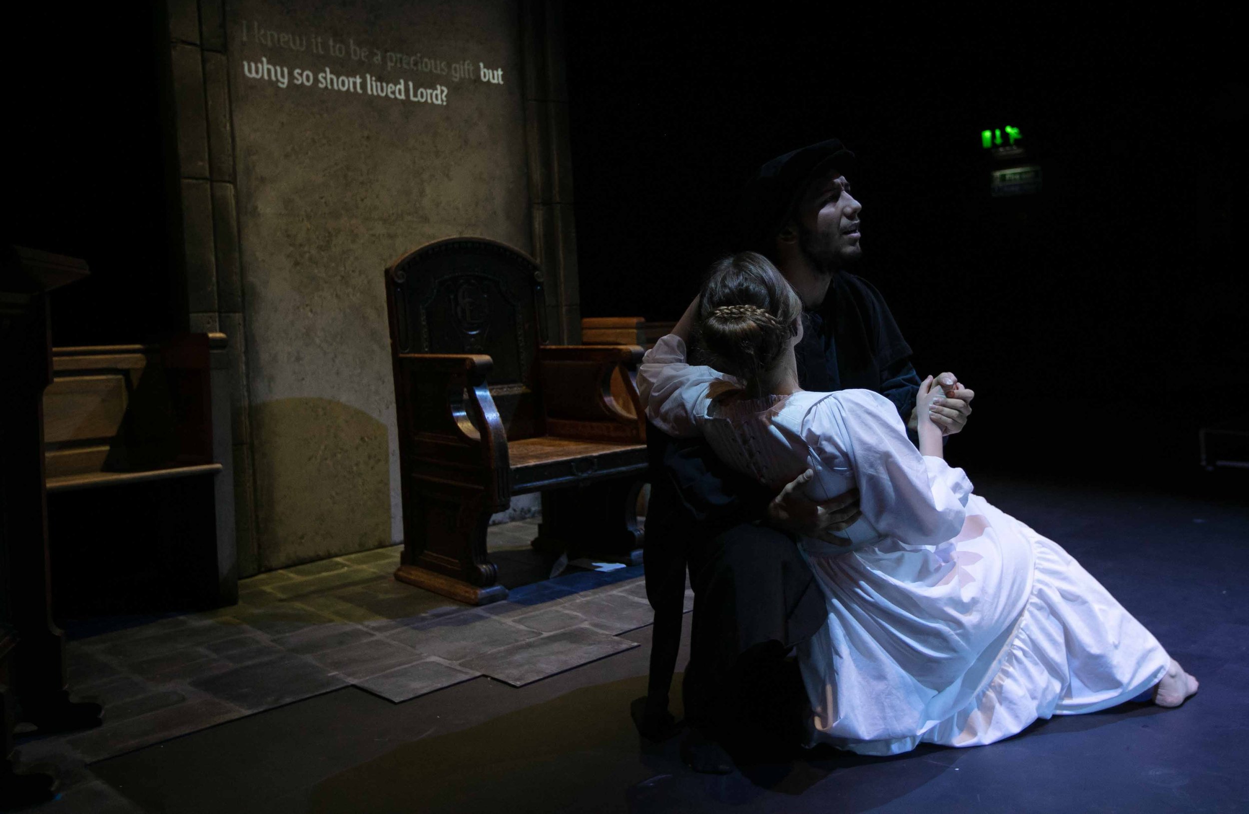 Gabriel Baicu as Knox cradles Claire Wetherall as Mary beside a stone arch bearing projected captions.