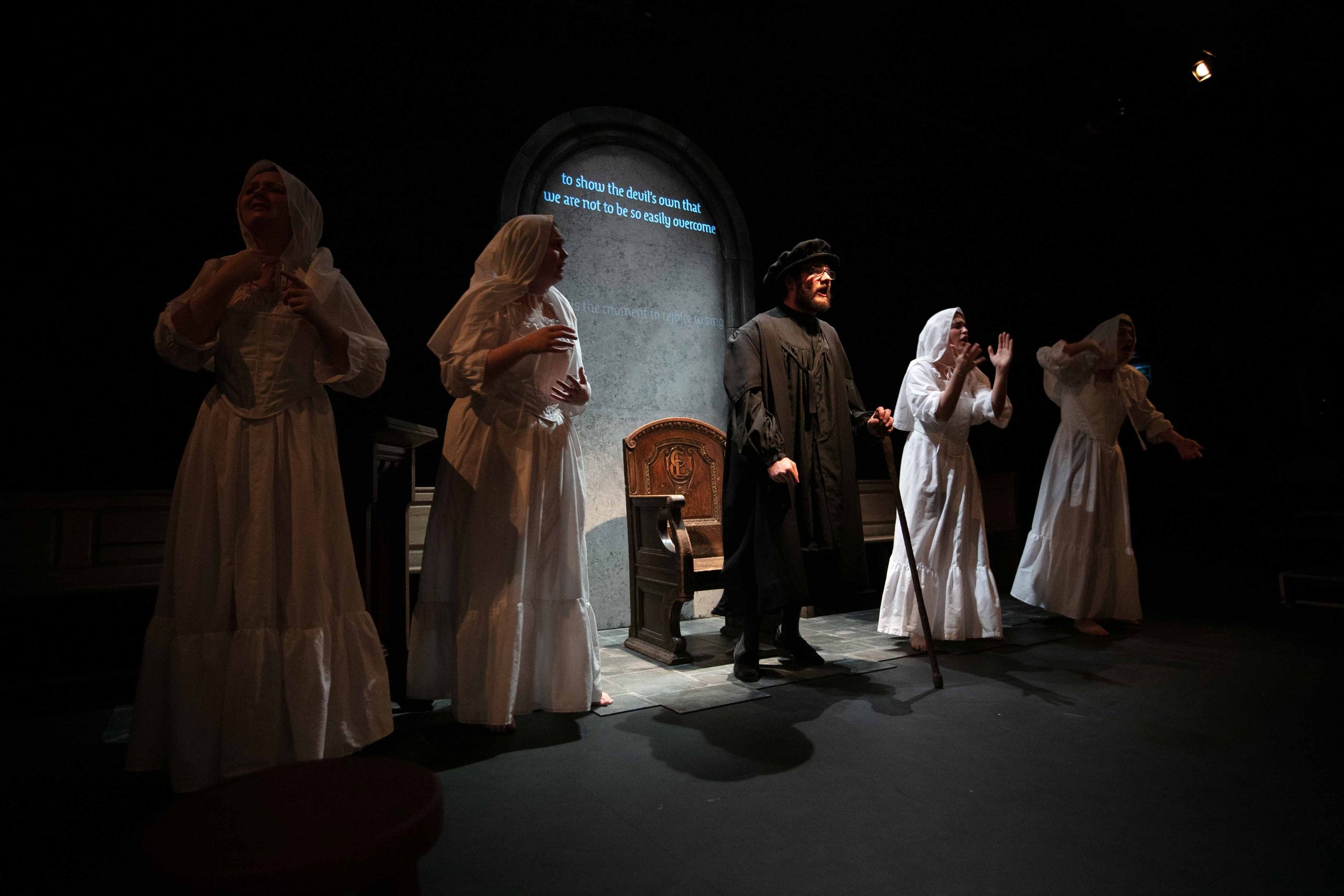 Neil Shand as Knox in black robes and cap among women in white, blue captions projected on the arch above.