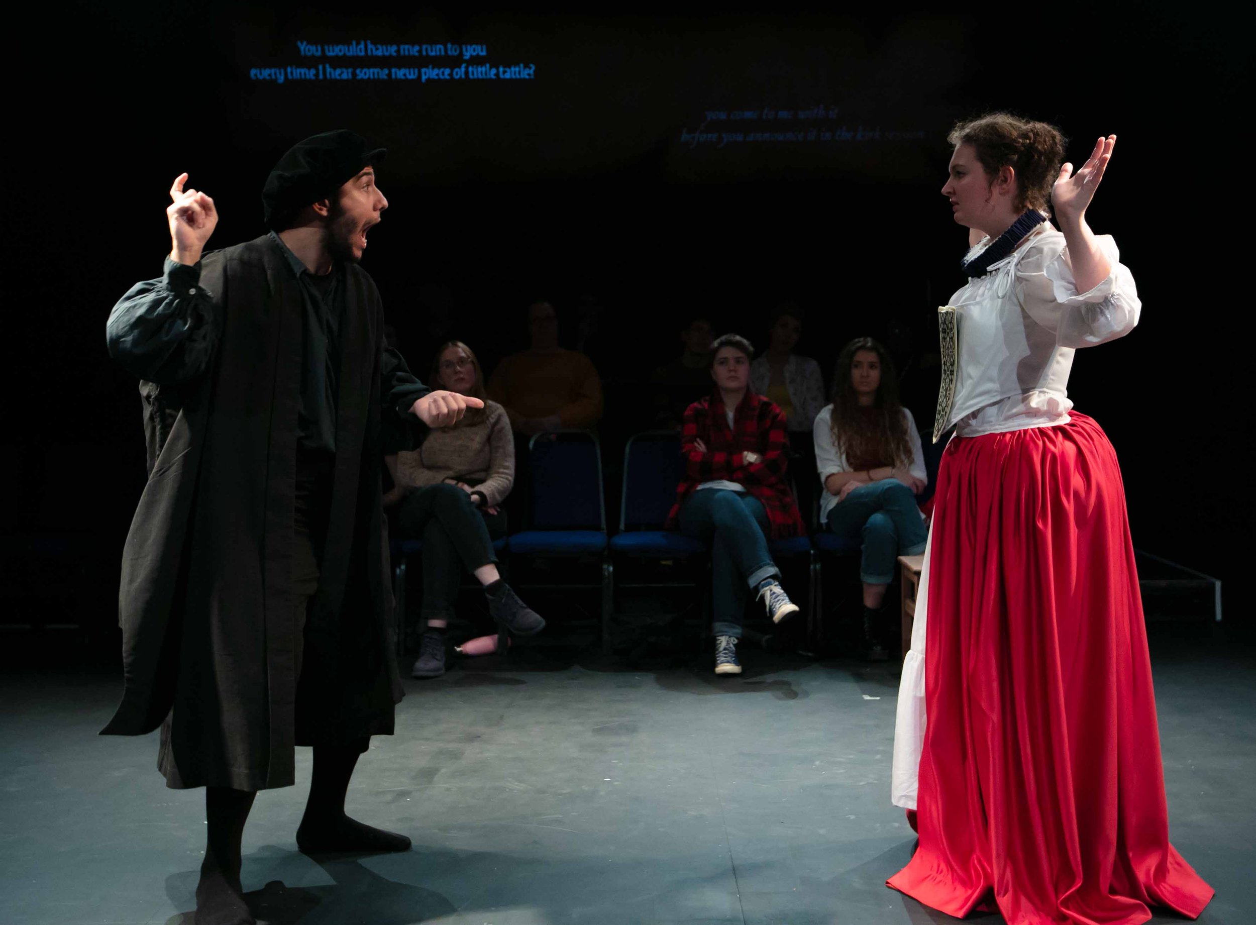 Gabriel Baicu as Knox gestures emphatically at Amy Helena as Mary, projected captions glowing overhead.