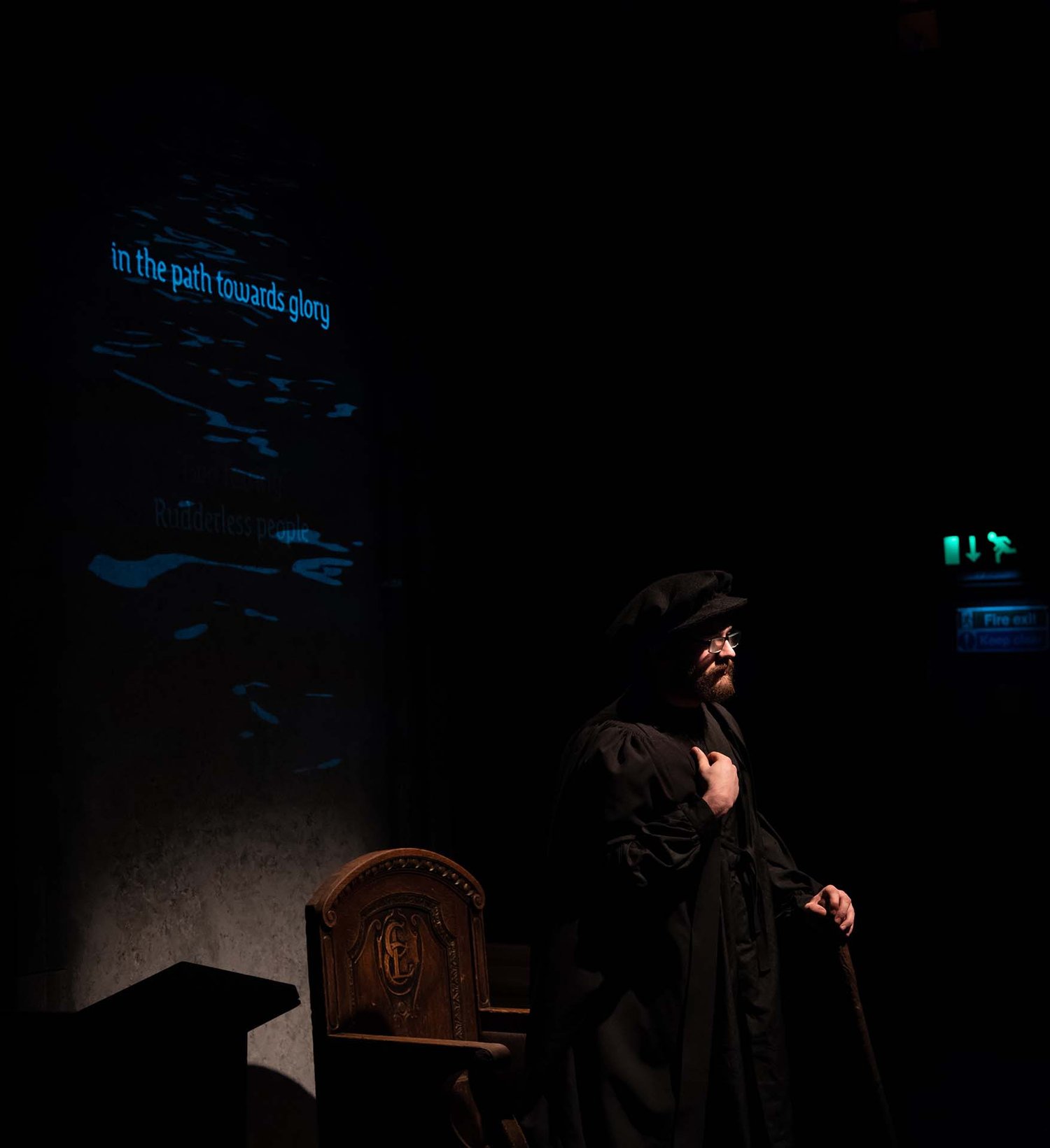 Neil Shand as Knox in half-light beside a rippling blue projection reading 'in the path towards glory'.