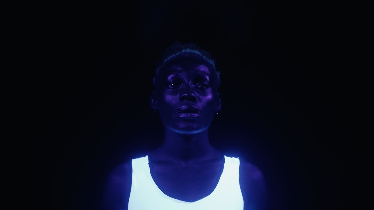 Film still from Grin: Divine Tasinda in a glowing white vest, her face lit violet against darkness