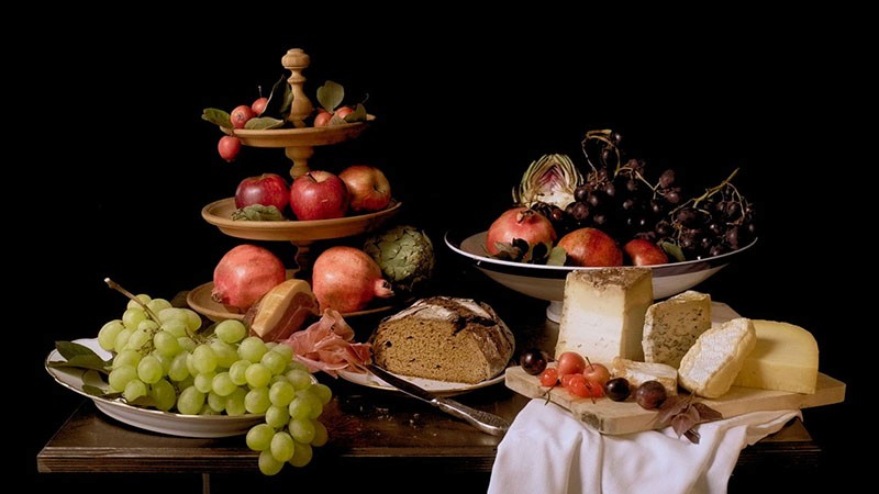Baroque-style still life of grapes, pomegranates, bread and cheeses on a table against black