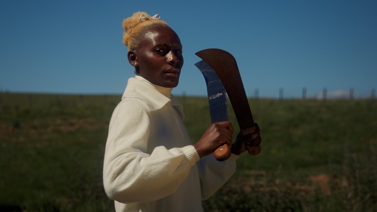 Divine Tasinda holds two machetes aloft in a green field; film still from Lagareh