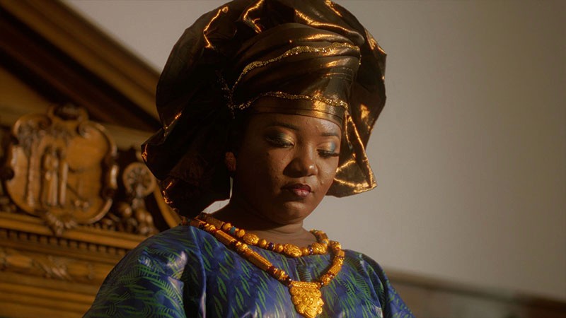 Kumba Kuyateh in a shimmering gold headwrap and gold necklace, eyes lowered; film still from Lagareh