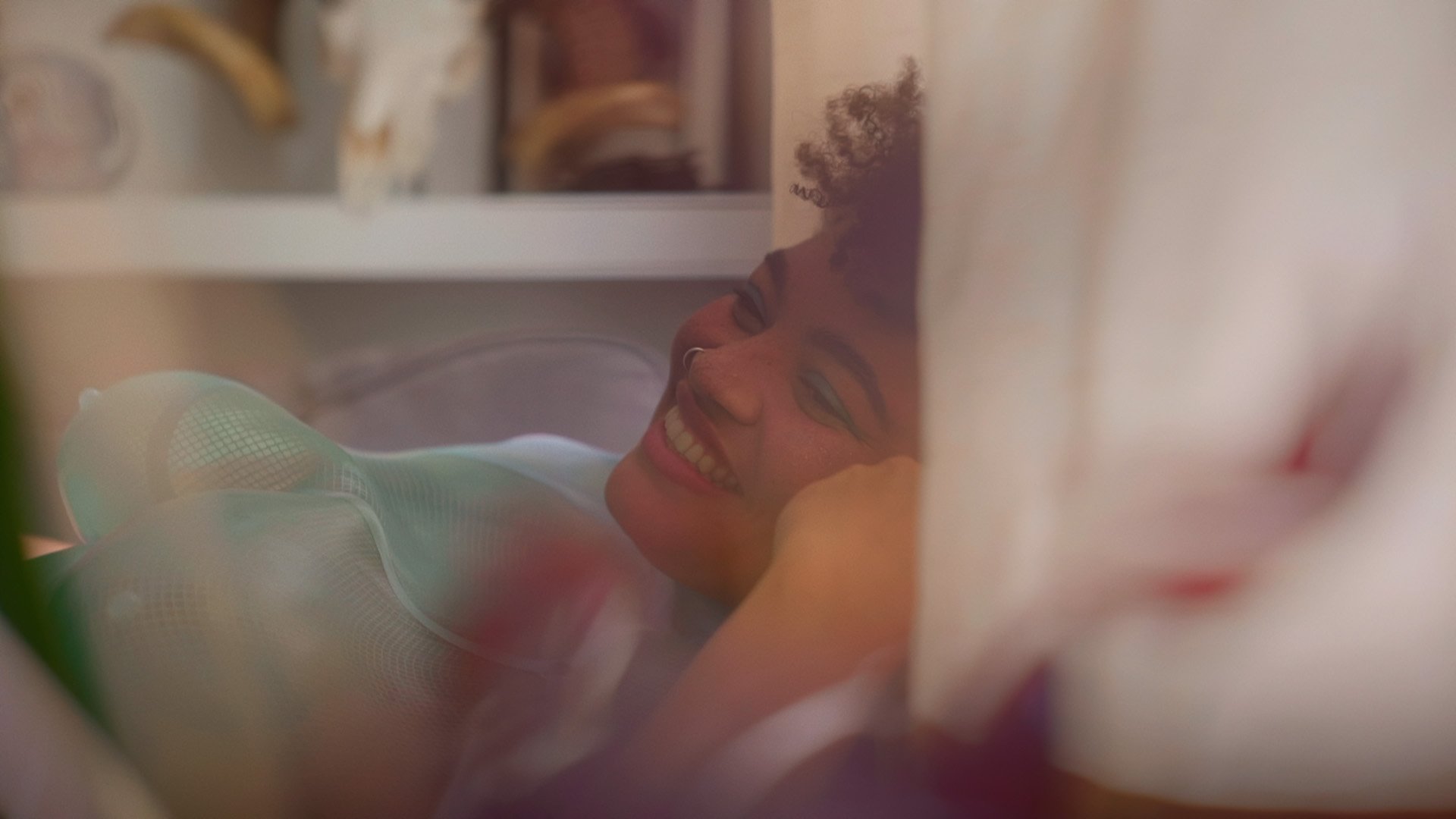 Ama at home, smiling softly behind a gauzy curtain; film still from Lagareh