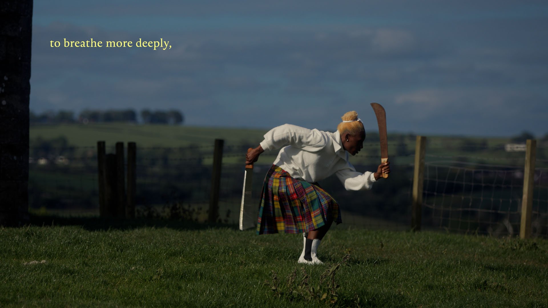 Divine Tasinda performs in a sunlit field, crouching with two machetes in a tartan skirt; film still from Lagareh