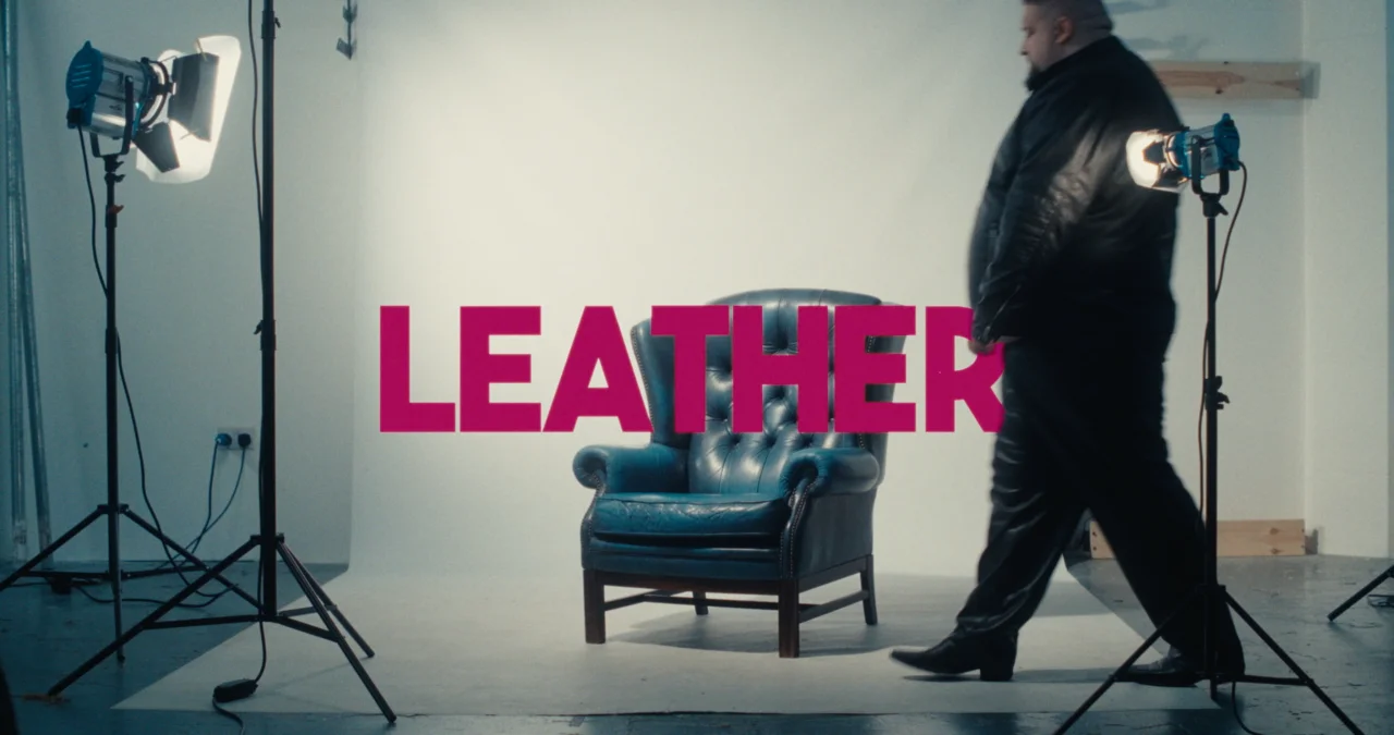 Music video title card: pink LEATHER lettering over a blue armchair on a white studio set with film lights