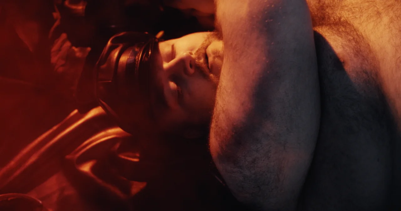 Music video still: red-lit close-up of a man in a leather cap lying back, bare arm raised