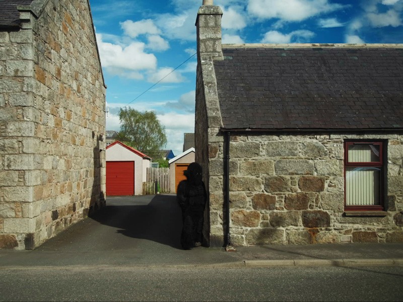 Film still: the shadowy Monster stands against a stone cottage beside an alley with a red garage door