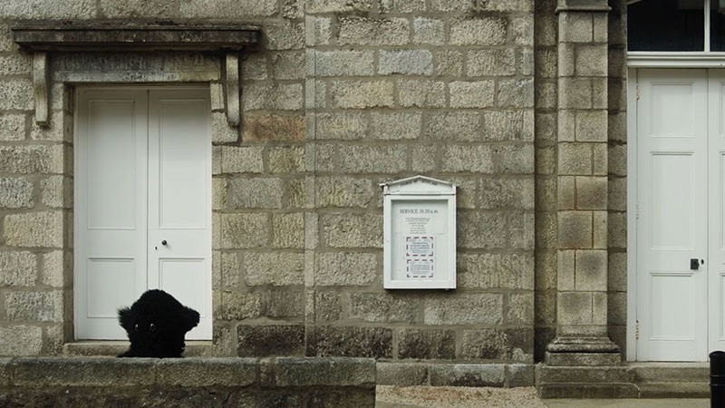 Film still: the furry Monster huddles low in a doorway of a granite building with white panelled doors
