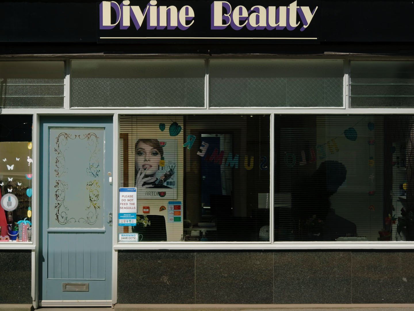 Film still: sunlit Divine Beauty salon shopfront with a pale blue door and a dark silhouette in the window